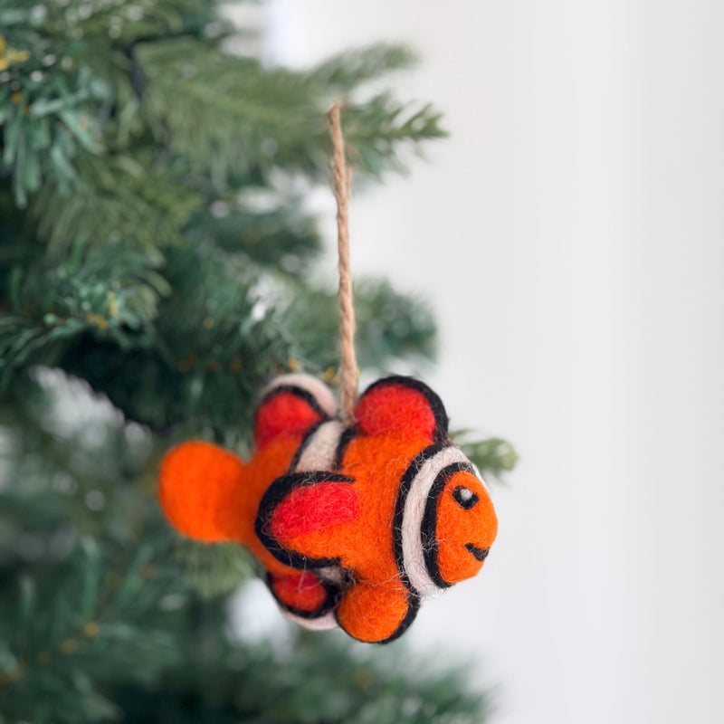 A playful felt clownfish ornament hangs from a green Christmas tree. This mini wool felt clownfish ornament is primarily orange with distinctive white and black stripes, capturing the vibrant look of a clownfish. A jute string is attached to the top for easy hanging, adding a cheerful, festive touch to the tree.