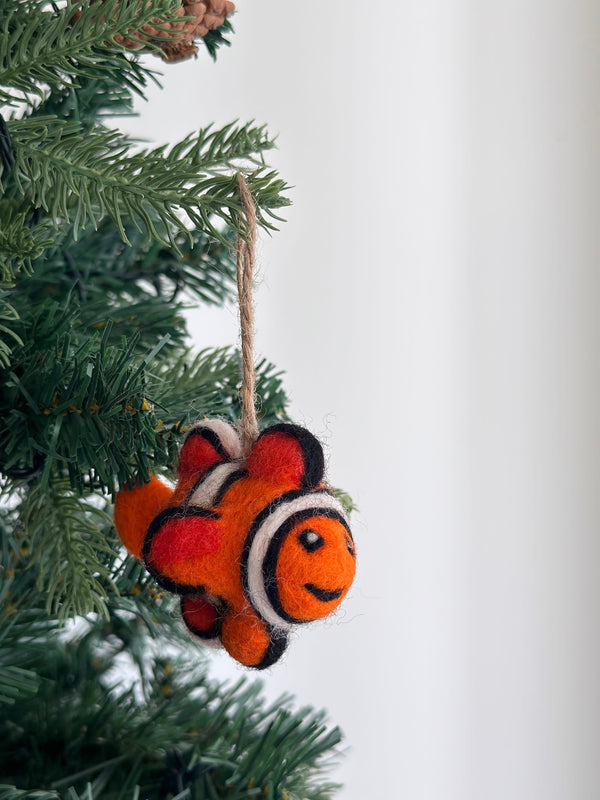 A mini clownfish wool felt ornament hangs from a pine tree branch, set against a soft white background, creating a cheerful, festive atmosphere. This vibrant orange clownfish ornament features distinct white and black stripes encircling its body. Crafted from soft wool felt for a tactile, handcrafted look, it includes a jute string for easy hanging.