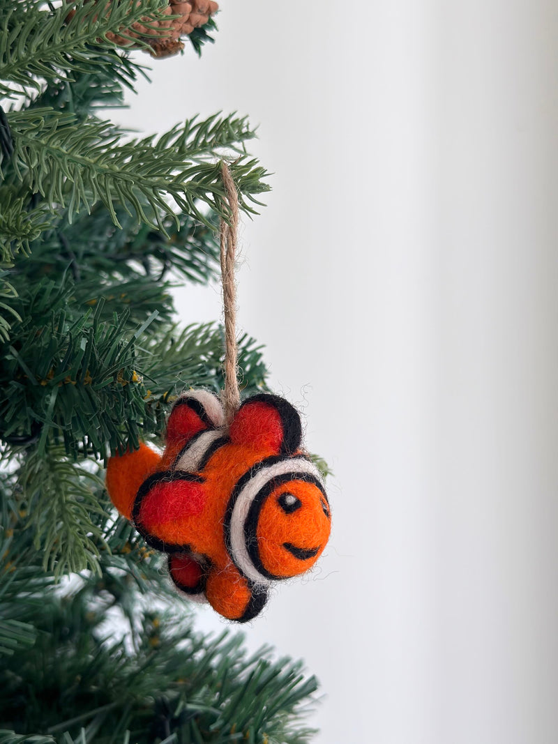 A mini clownfish wool felt ornament hangs from a pine tree branch, set against a soft white background, creating a cheerful, festive atmosphere. This vibrant orange clownfish ornament features distinct white and black stripes encircling its body. Crafted from soft wool felt for a tactile, handcrafted look, it includes a jute string for easy hanging.