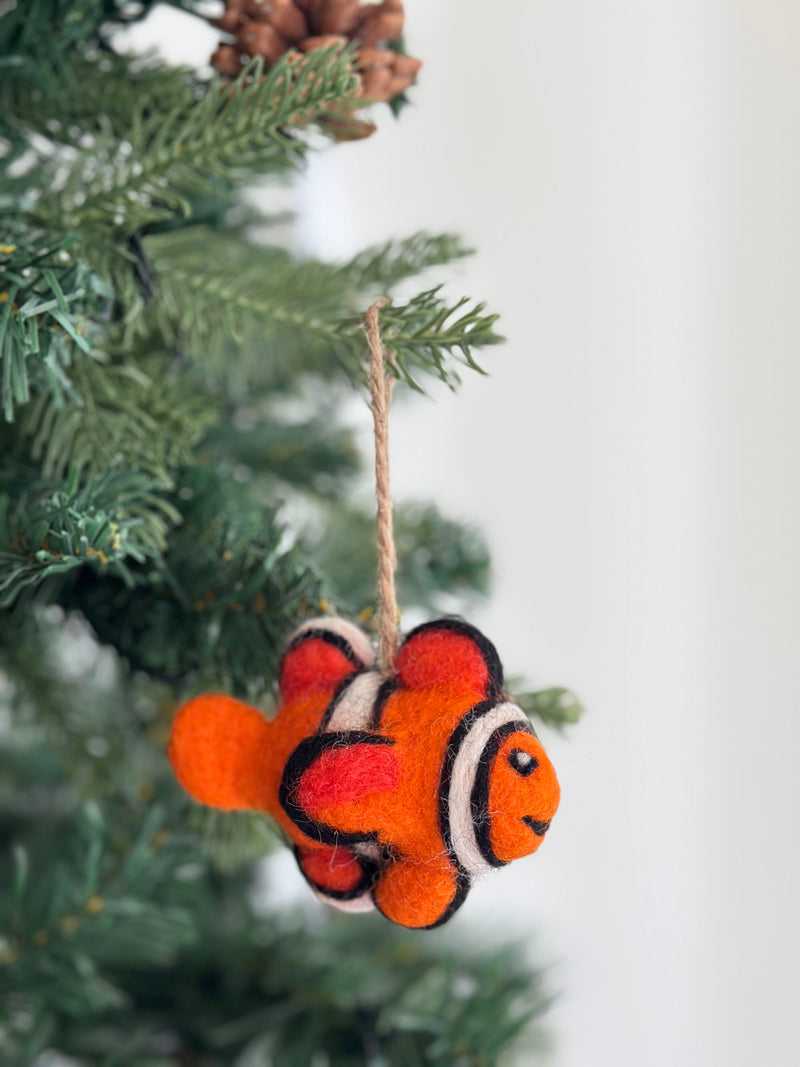 A vibrant wool felt mini clownfish ornament hangs from a Christmas tree branch against a white background. This handcrafted ornament, made from soft sheep wool felt, features bright orange, white, and black stripes, and a cheerful smile. A jute string is attached for easy hanging, adding a whimsical touch to festive decor.