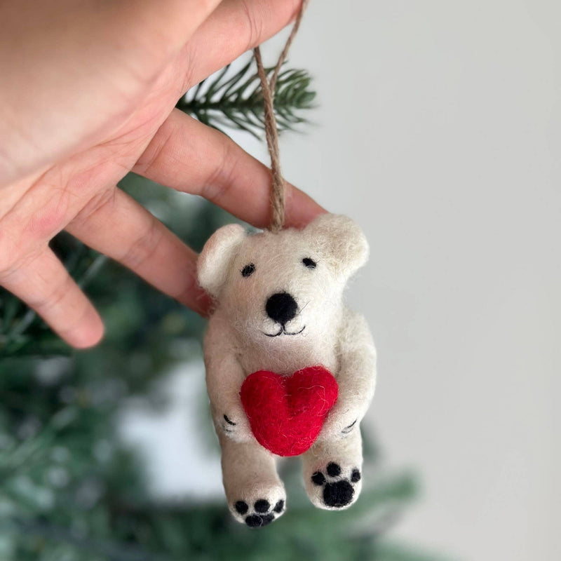 A handcrafted felt polar bear ornament is gently held by a female hand, positioned in front of a blurred Christmas tree. This charming mini polar bear, made of soft white wool, features a black stitched nose, mouth, and felt paws. It sweetly holds a bright red felt heart. A jute string on top completes this festive decoration.