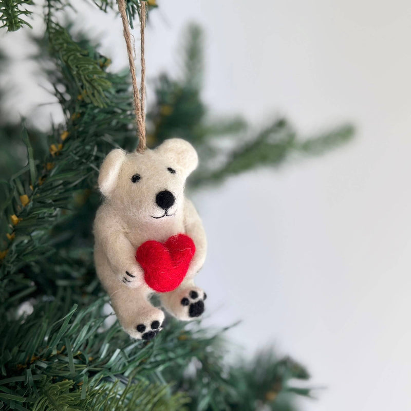 A handcrafted Mini Polar Bear Holding Heart felt ornament hangs gracefully on a Christmas tree. This charming white wool felt polar bear features a black nose, tiny black eyes, and feet, holding a bright red heart. A jute string allows this unique Christmas ornament to add a joyful, festive touch to any tree.