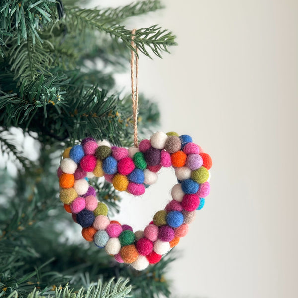 A wool felt Mini Pompom Heart Wreath ornament hangs from a Christmas tree branch against a soft white background. This petite, heart-shaped felt ornament is adorned with vibrant pink, red, white, blue, and orange mini pompoms, with a jute string loop at the top for hanging.