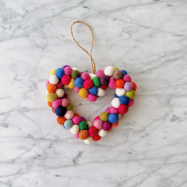A colorful wool felt mini pompom heart wreath ornament rests on a light, marbled surface. This handcrafted ornament features a petite heart shape, tightly packed with numerous mini pompoms in vibrant shades of white, pink, blue, red, green, orange, and yellow. A jute string is attached at the top for hanging, adding a playful and cheerful touch.