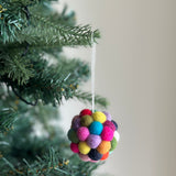 A colorful felt pompom ornament hangs from a green Christmas tree branch, creating a joyful, festive atmosphere. This mini felt ornament is spherical, crafted from numerous small, round felt balls in a rainbow of colors including red, yellow, green, blue, purple, pink, and orange. A thin white cotton string allows it to hang from the branch.