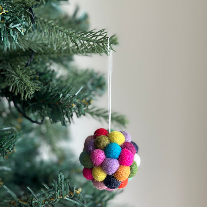 A colorful felt pompom ornament hangs from a green Christmas tree branch, creating a joyful, festive atmosphere. This mini felt ornament is spherical, crafted from numerous small, round felt balls in a rainbow of colors including red, yellow, green, blue, purple, pink, and orange. A thin white cotton string allows it to hang from the branch.