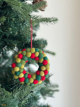 A wool felt mini pompom heart wreath ornament hangs from a Christmas tree branch. This handmade felt ornament features a petite heart shape adorned with green, yellow, and red mini pompoms, suspended by a red cotton string. The close-up shot highlights its cozy texture against the blurred tree background.
