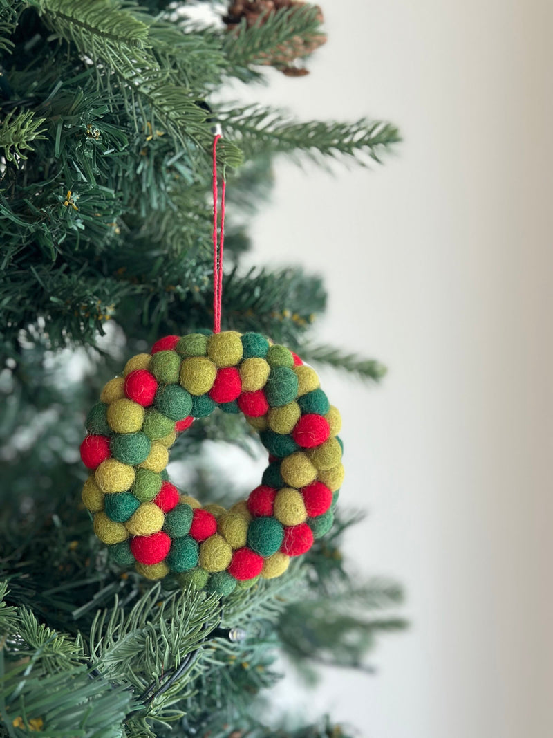 A wool felt mini pompom heart wreath ornament hangs from a Christmas tree branch. This handmade felt ornament features a petite heart shape adorned with green, yellow, and red mini pompoms, suspended by a red cotton string. The close-up shot highlights its cozy texture against the blurred tree background.