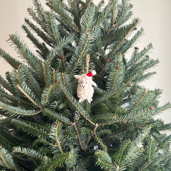 A handmade wool felt mini sheep ornament hangs on a festive green Christmas tree. This cream-colored sheep features a fluffy body, a stitched black smiling mouth, and a red and white Santa hat jauntily placed on its head. It adds a whimsical touch to the holiday decor.