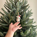 A charming mini sheep felt ornament, held gently by a hand, hangs from the branch of a lush Christmas tree. This delightful, cream-colored wool felt sheep features a fluffy texture, perky ears, and a stitched 'Y' for a nose. It wears a tiny red and white Christmas hat and hangs from a jute string, adding a festive touch to the holiday decor.