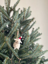 A mini sheep felt ornament with a Christmas hat hangs on a festive Christmas tree. This cream-colored, handcrafted wool felt ornament has a textured, needle-felted finish. It features a red and white Christmas hat, black stitched facial details, and a jute string for hanging, adding to the joyful atmosphere.