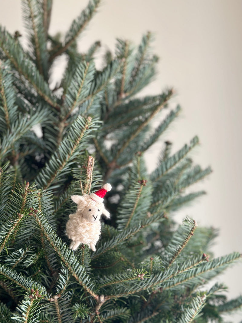 A mini sheep felt ornament with a Christmas hat hangs on a festive Christmas tree. This cream-colored, handcrafted wool felt ornament has a textured, needle-felted finish. It features a red and white Christmas hat, black stitched facial details, and a jute string for hanging, adding to the joyful atmosphere.