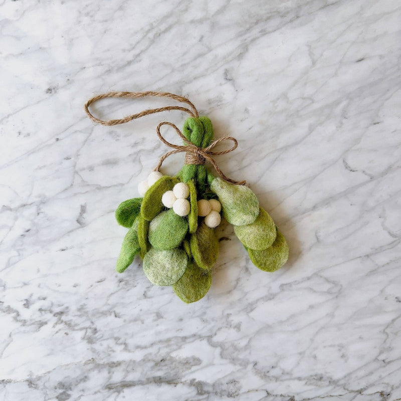 A handcrafted felt mistletoe sprig Christmas ornament is presented on a softly lit, light marble surface. This unique Christmas ornament features distinctive tie-dye felt leaves and clusters of white felt berries. A rustic jute string forms a bow at the top, ready for hanging.