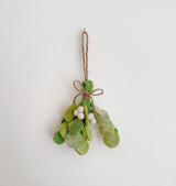 A felt mistletoe sprig ornament is displayed against a plain white wall. This unique Christmas ornament features tie-dye felt leaves in varying shades of green, creating a textured look. A jute string with a decorative bow at the top is attached for hanging.