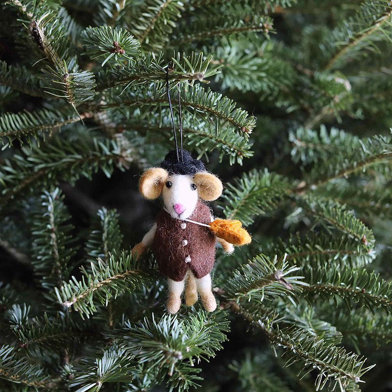 A handmade felt mouse boy ornament hangs on a fresh green Christmas tree. This needle felted wool mouse features a white face, tan ears, and a pink stitched nose. Dressed in a brown vest with buttons and a black hat, it playfully holds a yellow leaf. A black string is attached to the hat for easy hanging.