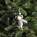 A charming needle-felted felt mouse ornament, wearing a gray sweater with a red snowflake, matching hat, and scarf, hangs playfully on a lush green Christmas tree branch. The adorable mouse features tan ears, hands, and feet, and a cream-colored face, with a white string for easy hanging.