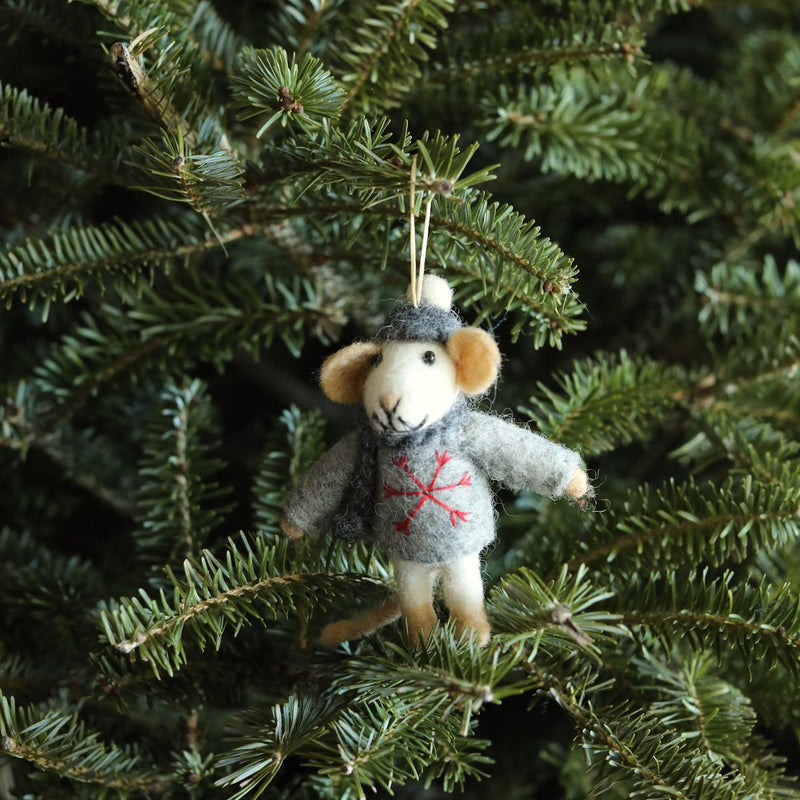 A charming needle-felted felt mouse ornament, wearing a gray sweater with a red snowflake, matching hat, and scarf, hangs playfully on a lush green Christmas tree branch. The adorable mouse features tan ears, hands, and feet, and a cream-colored face, with a white string for easy hanging.