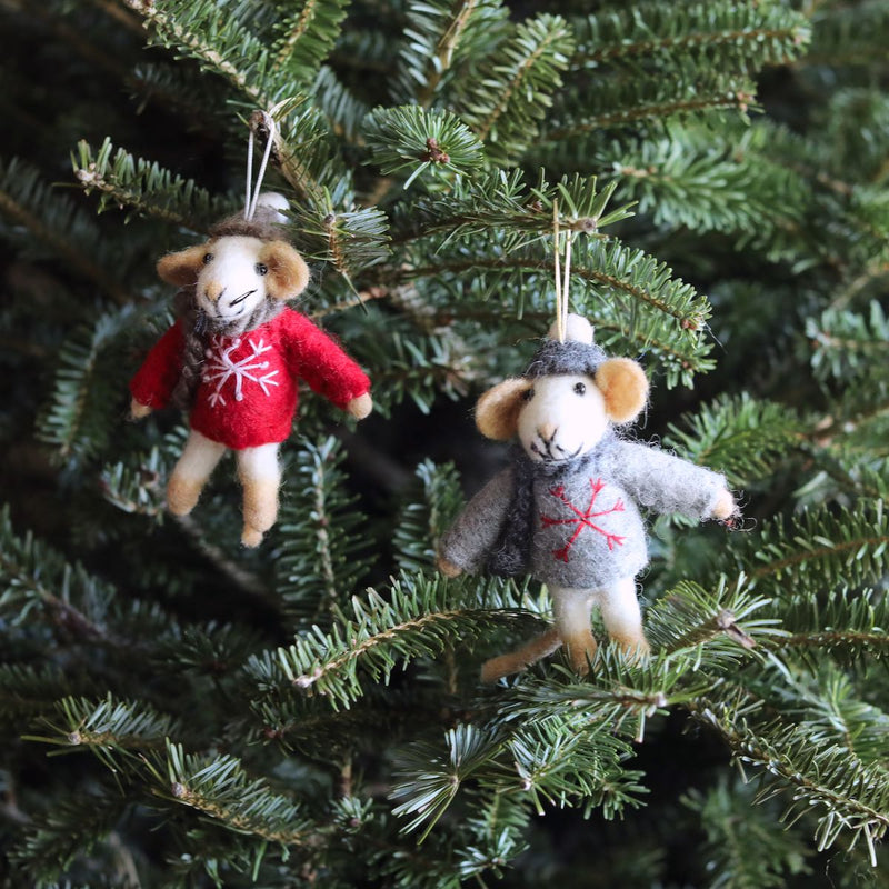 A needle felted mouse Christmas ornament is suspended from a branch on a lush green Christmas tree. This charming handmade wool felt mouse wears a gray sweater adorned with a red snowflake, complemented by tan ears and a tan tail. It adds a whimsical and joyful touch to the holiday decor.
