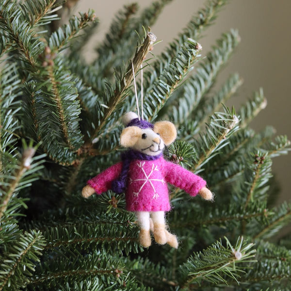 A charming felt mouse Christmas ornament hangs among the vibrant green needles of a Christmas tree. This 3D needle felted mouse has a cream-colored head, round ears, and a stitched smile. Dressed in a purple knit cap and matching scarf, it also wears a bright pink sweater with a white snowflake. A white string is attached for easy hanging.