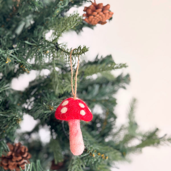 A cheerful felt mushroom ornament hangs on a green Christmas tree adorned with green needles and pine cones. The felt mushroom ornament features a bright red cap with white dots and a pale pink stem, with a jute string attached for hanging.