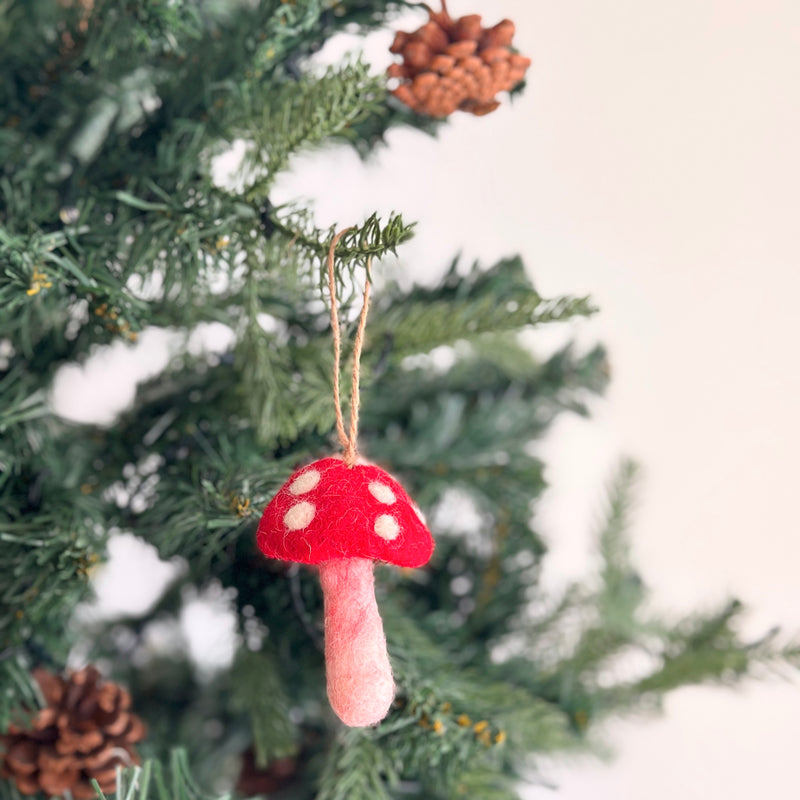 A cheerful felt mushroom ornament hangs on a green Christmas tree adorned with green needles and pine cones. The felt mushroom ornament features a bright red cap with white dots and a pale pink stem, with a jute string attached for hanging.