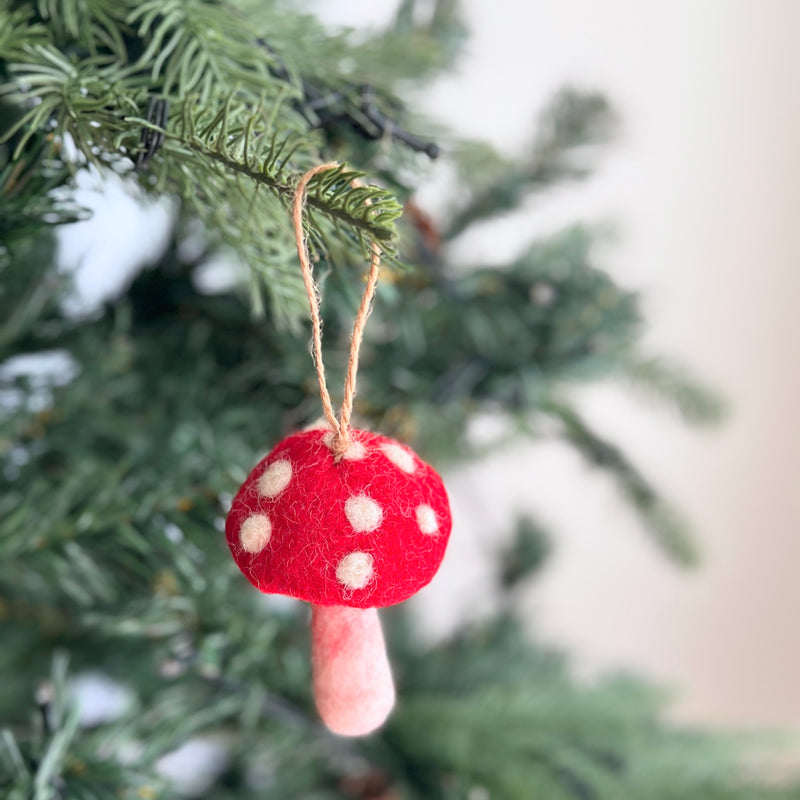A charming felt mushroom ornament hangs on a lush green Christmas tree, evoking a joyful and celebratory mood. This handcrafted felt Christmas ornament features a vibrant red cap with white dots and a light pink stem, suspended by a jute string. It adds a whimsical touch to holiday decor.