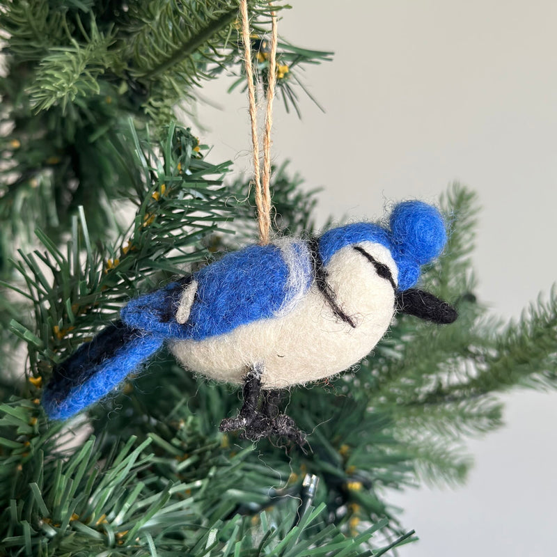 A needle felted blue jay ornament hangs from a jute string on a Christmas tree. The blue jay features vibrant blue and white plumage, detailed with black felt accents, adding a festive touch of nature to the holiday decor.