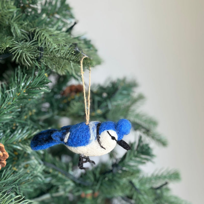A needle felted blue jay ornament hangs gracefully on a green pine tree branch, evoking a natural and joyful atmosphere. This felt blue jay is predominantly blue with white accents on its face and underbelly, a black beak, and delicate felted details forming its legs. A jute string is attached for hanging.