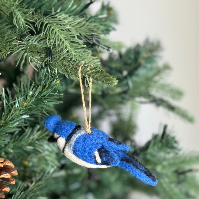A needle felted blue jay felt ornament, featuring a vibrant blue body with black and white markings and a fluffy crest, hangs by a jute string from a green branch of a decorated Christmas tree.