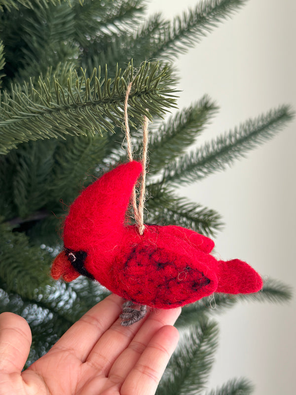 A vibrant red needle felted cardinal ornament, suspended by a jute string, is gently held by a female hand against a blurred Christmas tree backdrop. This handmade felt cardinal ornament features black accents around its face and on its back, evoking warmth and tradition for holiday decorating.