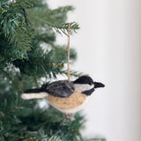 A charming needle felted chickadee ornament hangs gracefully on a Christmas tree branch, with a blurred backdrop of green pine needles, creating a natural and festive mood. This unique handmade felt ornament features a predominantly black head and back, gray and white patterned wings, and a tan body. A jute string is attached for easy hanging.