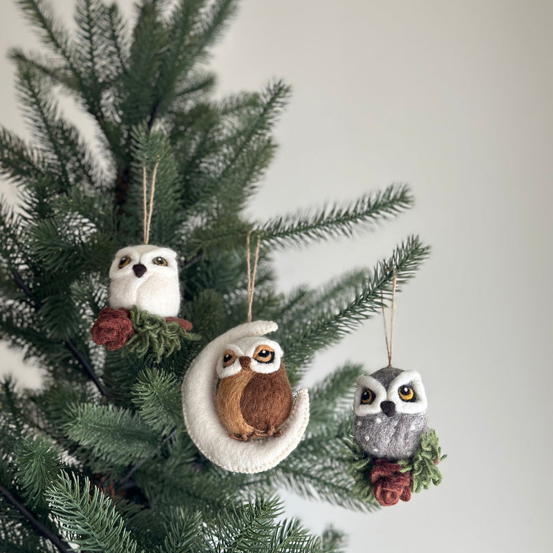 Three needle felted owl ornaments are displayed hanging on a Christmas tree, creating a festive and charming scene. These unique Christmas tree ornaments include a white owl perched on a cypress tree, a brown owl nestled in a white moon, and a gray owl atop another cypress tree. Each wood-themed, handcrafted felt owl ornament is detailed with small eyes and includes a jute string for easy hanging.
