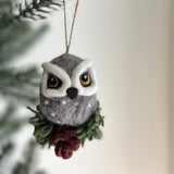 A handcrafted needle felted owl ornament is suspended from a Christmas tree branch, creating a cozy, festive atmosphere. This grey owl ornament features white accents around its yellow eyes with black pupils, a dark brown beak, and subtle grey spots on its chest. The owl perches on a cypress tree base with green felt foliage and a brown felt pine cone.