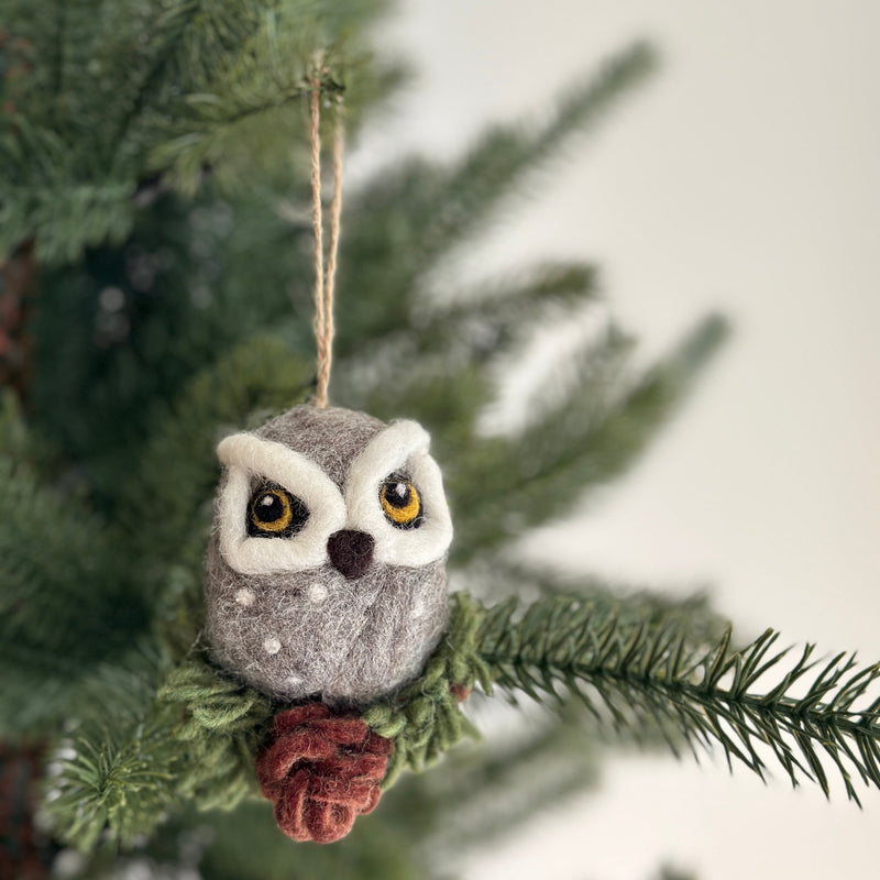 A needle felted owl ornament hangs on a green Christmas tree branch, evoking a sense of handmade charm. The grey owl features white accents around its eyes and small white spots on its body. A green cypress tree branch element is visible below the owl, and a jute string is attached to its top for hanging.