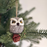 A grey needle felted owl ornament hangs from a pine branch of a Christmas tree. This charming handcrafted felt owl features prominent yellow eyes, a small brown beak, and white accents, perched on green felted cypress leaves. A jute string extends from the top, allowing it to hang as a Christmas tree ornament.