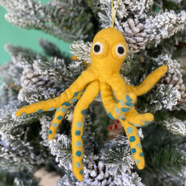 A yellow felt octopus ornament hangs on a snow-flocked Christmas tree. This playful octopus features eight legs extending from its body and two big eyes. A yellow cotton string is attached at the top for easy hanging.