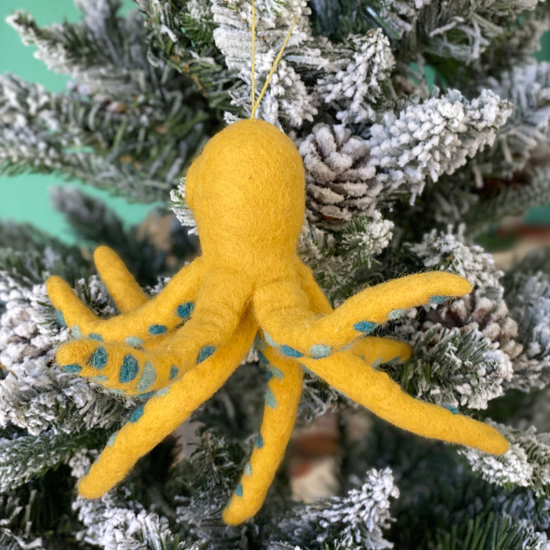 A yellow octopus felt ornament hangs joyfully on a snow-flocked Christmas tree. This whimsical wool felt octopus ornament features a rounded head and eight posable wire legs. A matching yellow cotton string allows it to blend seamlessly with the festive branches and pine cones.