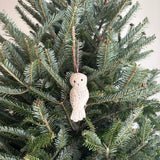 A needle felted owl ornament rests among the lush green needles of a Christmas tree. This off-white wool felt owl features a simple, stylized form with a round head, beak, and eyes. Subtle stitched details suggest feathers, and a red and white cotton string is attached for hanging, adding a touch of woodland charm.
