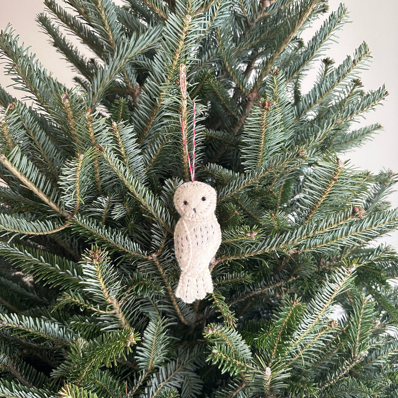 A needle felted owl ornament rests among the lush green needles of a Christmas tree. This off-white wool felt owl features a simple, stylized form with a round head, beak, and eyes. Subtle stitched details suggest feathers, and a red and white cotton string is attached for hanging, adding a touch of woodland charm.