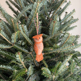 A needle felted owl ornament hangs on a vibrant green Christmas tree. The owl features soft, two-tone brown felt with delicate stitch details. A red and white cotton string is attached, ready for display.