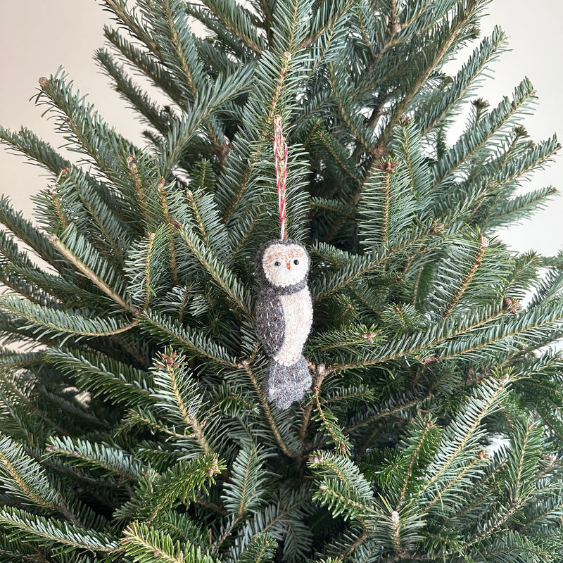 A handmade wool felt owl ornament from the 'Owl Family' collection hangs gracefully from a Christmas tree branch. This needle-felted owl features a white face, gray body, small round eyes, and a small beak.