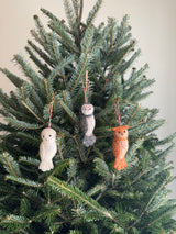 Three wool felt owl ornaments hang on the green needles of a Christmas tree. These unique Christmas owl ornaments include one cream with black spots, one gray and cream, and one light orange, each with a cotton string for hanging.