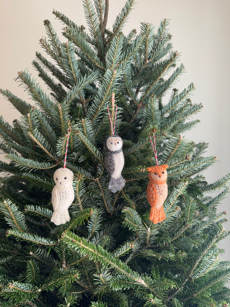 Three wool felt owl ornaments hang on the green needles of a Christmas tree. These unique Christmas owl ornaments include one cream with black spots, one gray and cream, and one light orange, each with a cotton string for hanging.