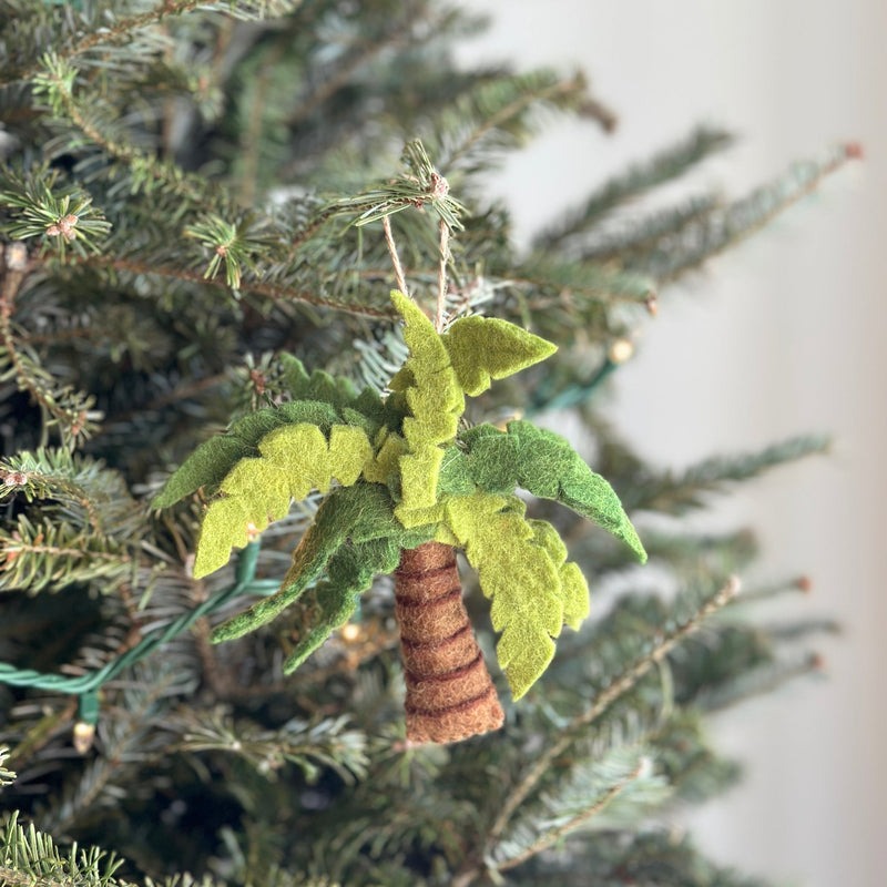 A handcrafted wool felt palm tree ornament hangs prominently on a Christmas tree, with blurred string lights in the background. This unique Christmas ornament features a detailed brown trunk with horizontal stripes and layered green fronds in varying shades, bringing a cheerful, tropical touch to the holiday decor.