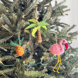 A handmade felt palm tree ornament hangs on a Christmas tree. The unique Christmas ornament, crafted from sheep's wool, features a brown trunk and green leaves, suspended by a jute string. Small white lights adorn the festive tree.