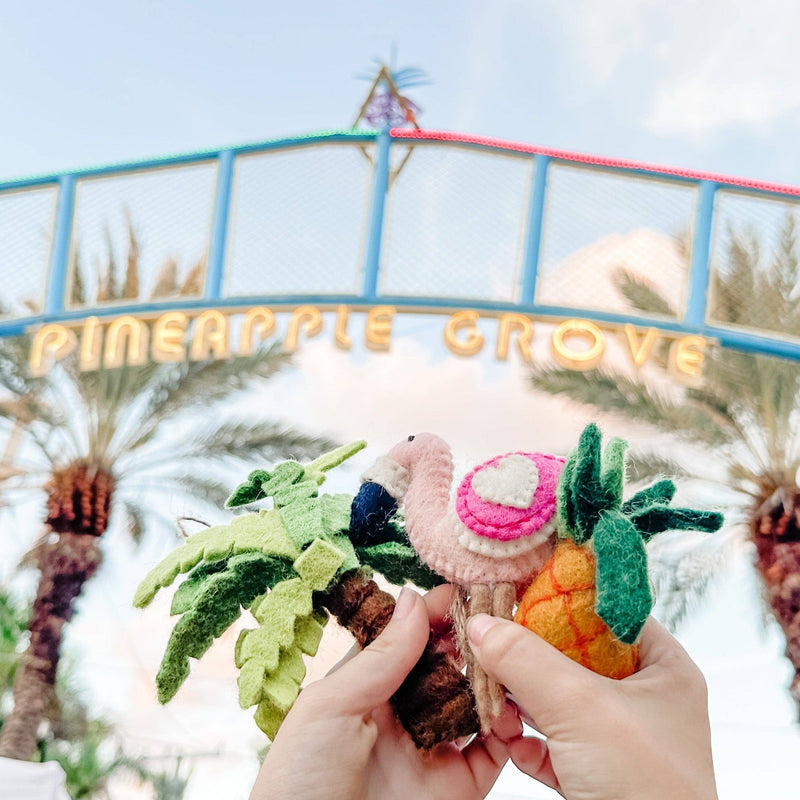 A vibrant, handmade felt palm tree ornament is delicately held by a female hand, set against a bright, airy backdrop featuring palm trees and a "Pineapple Grove" sign that evokes a cheerful, tropical atmosphere. This unique Christmas ornament showcases detailed green fronds and a brown trunk, complete with a jute string for easy hanging.