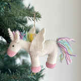 A pastel unicorn felt ornament hangs playfully on a Christmas tree, positioned against an off-white wall. This whimsical wool felt unicorn features a soft white body, pink snout, and pink hooves. Its mane and tail boast pastel yellow, green, blue, and purple strands, complemented by a spiraled white horn and white felt wings, with a jute string attached for hanging.