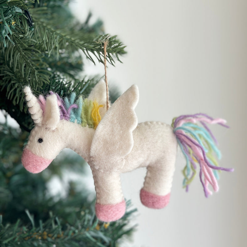 A pastel unicorn felt ornament hangs playfully on a Christmas tree, positioned against an off-white wall. This whimsical wool felt unicorn features a soft white body, pink snout, and pink hooves. Its mane and tail boast pastel yellow, green, blue, and purple strands, complemented by a spiraled white horn and white felt wings, with a jute string attached for hanging.