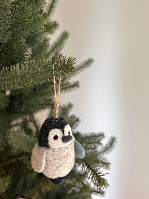 A wool felt penguin ornament hangs from a Christmas tree branch, adding a festive touch. The penguin features a black head and back, a beige belly, and grey wings, with black eyes and a beak. A jute string is attached to the top for easy hanging.