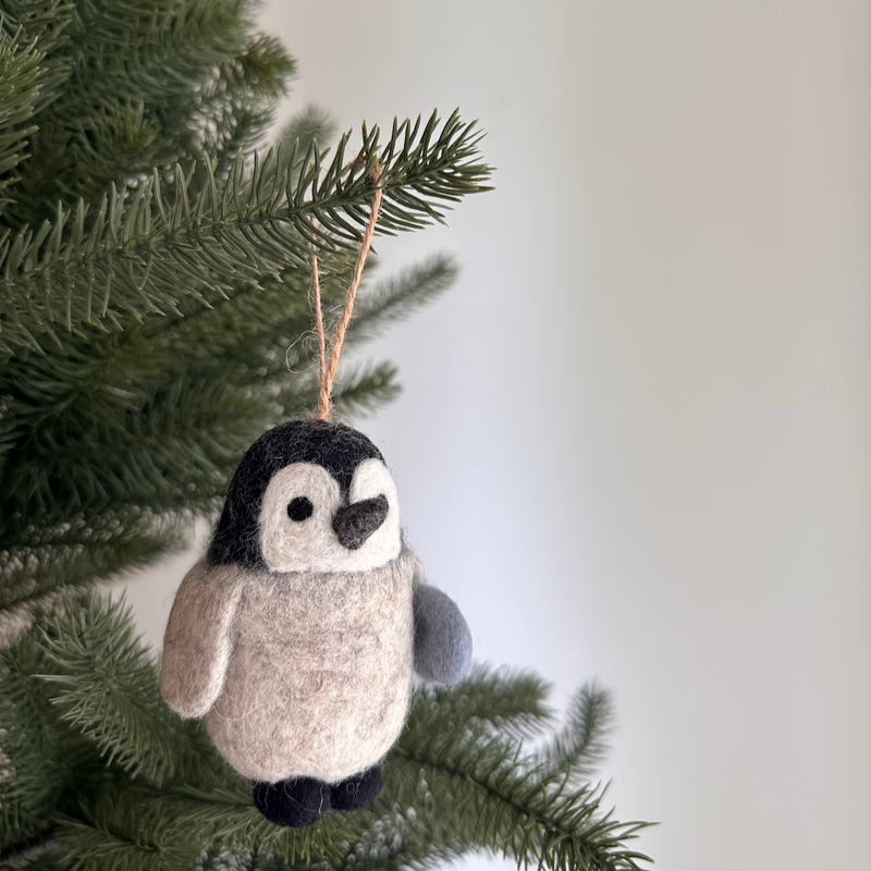 A cheerful wool felt penguin ornament, depicted holding a fish, hangs on a lush green Christmas tree. The charming penguin has a gray body, white face, and a black head, feet, and beak with black eyes. Its gray felted wings add texture, and a jute string is attached for easy hanging.